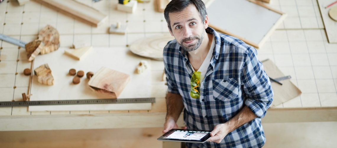 Content handsome middle-aged man with beard frowning forehead and holding tablet while leaning on table with wooden details and looking at camera in carpentry